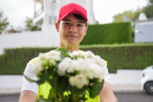 Smiling delivery man with a red cap offering a bouquet of white flowers outdoors in Portugal.